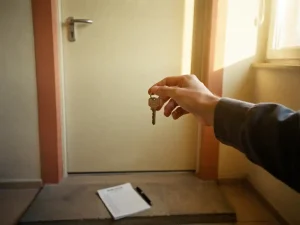 Hand van huurder met huissleutel voor pasgeverfde appartementsdeur, checklist en pen op de drempel, warm middaglicht.