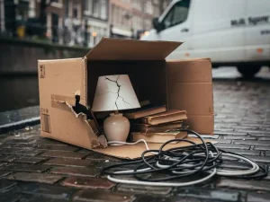 Opengebarsten verhuisdoos op Amsterdamse klinkers met gebarsten lamp, verspreide boeken en kabels, verhuiswagen op de achtergrond.