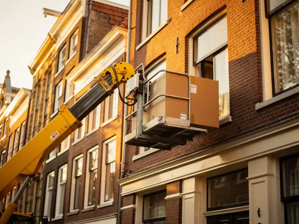Gele verhuislift naast gevel van Amsterdams grachtenpand met kartonnen verhuisdoos in de lucht, warme middagzon op bakstenen gevel.