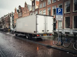 Witte verhuiswagen op smalle Amsterdamse gracht, parkeerontheffingsbord, historische gevels en fietsen op klinkers.