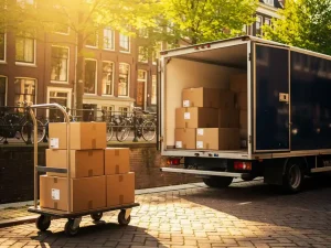 Verhuiswagen geparkeerd op een zonnige Amsterdamse gracht, met kartonnen dozen op een steekwagen naast bakstenen gevels.