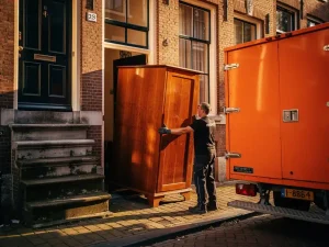 Verhuizer die een houten kledingkast door een smalle deuropening van een Amsterdams grachtenpand manoeuvreert, oranje verhuiswagen op kasseienstraat.