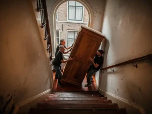Verhuizers tillen een grote houten kast door het steile trappenhuis van een Amsterdams grachtenpand, met bakstenen muren en boogvenster.