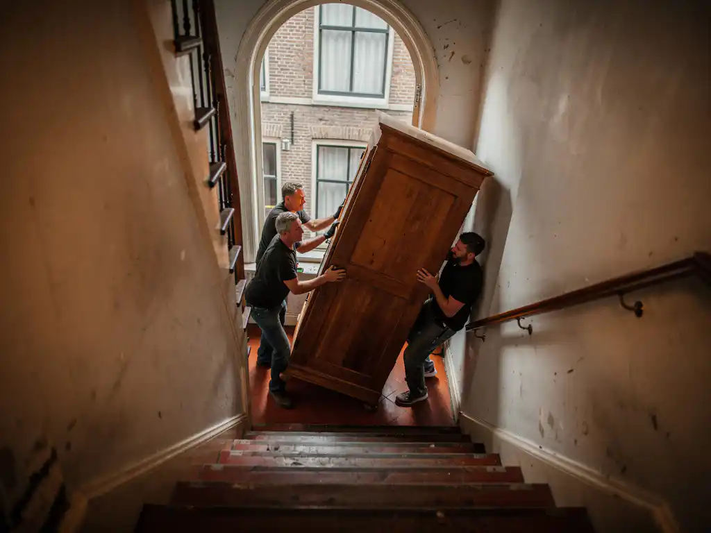 Verhuizers tillen een grote houten kast door het steile trappenhuis van een Amsterdams grachtenpand, met bakstenen muren en boogvenster.