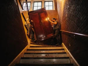 Verhuizers tillen een houten kledingkast door een steile, smalle trappenhuis van een Amsterdams grachtenpand in de Jordaan.