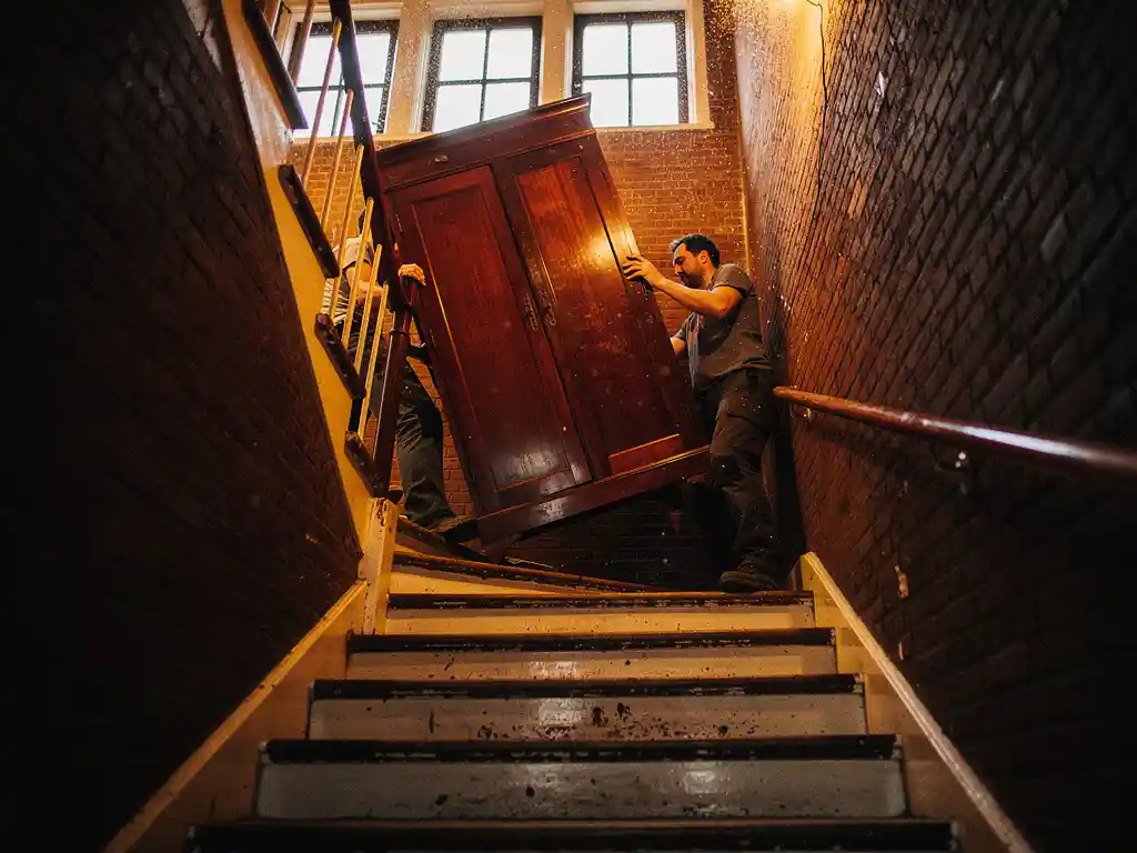 Verhuizers tillen een houten kledingkast door een steile, smalle trappenhuis van een Amsterdams grachtenpand in de Jordaan.