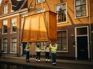 Houten kledingkast gehesen aan transportbanden langs een smalle Amsterdamse grachtenpandgevel, verhuizers begeleiden de lift in middaglicht.